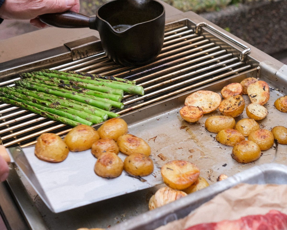 Person grilling vegetables and meat on a barbecue with a garden in the background